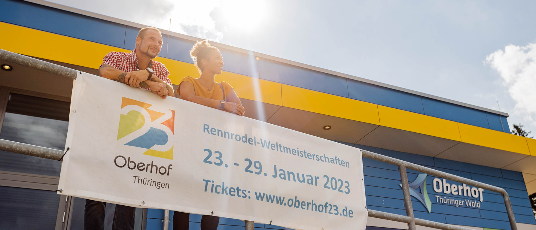 Alexander Rödiger und Mariama Jamanka lehnen an einer Brüstung. Vor ihnen hängt ein Plakat, dass die Rennrodel-WM in Oberhof vom 23.-29. Januar 2023 ankündigt.