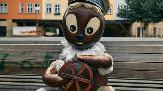 Die Pittiplatsch-Figur sitzt auf einer Holzbank und hält ein Rad in beiden Händen